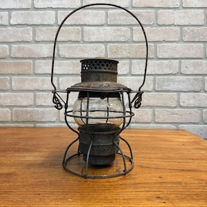 May include: An antique, rusty metal lantern with a clear glass globe. The lantern has a wire handle and a circular base. The glass globe is protected by a metal cage. The lantern sits on a wooden surface.