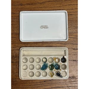 May include: A rectangular white metal watercolor paint box with a separate lid. The lid features the text F.W. DEVOE & CO. NEW YORK. The base contains twenty circular wells for paint, with some residual blue, green, yellow, and black pigment.