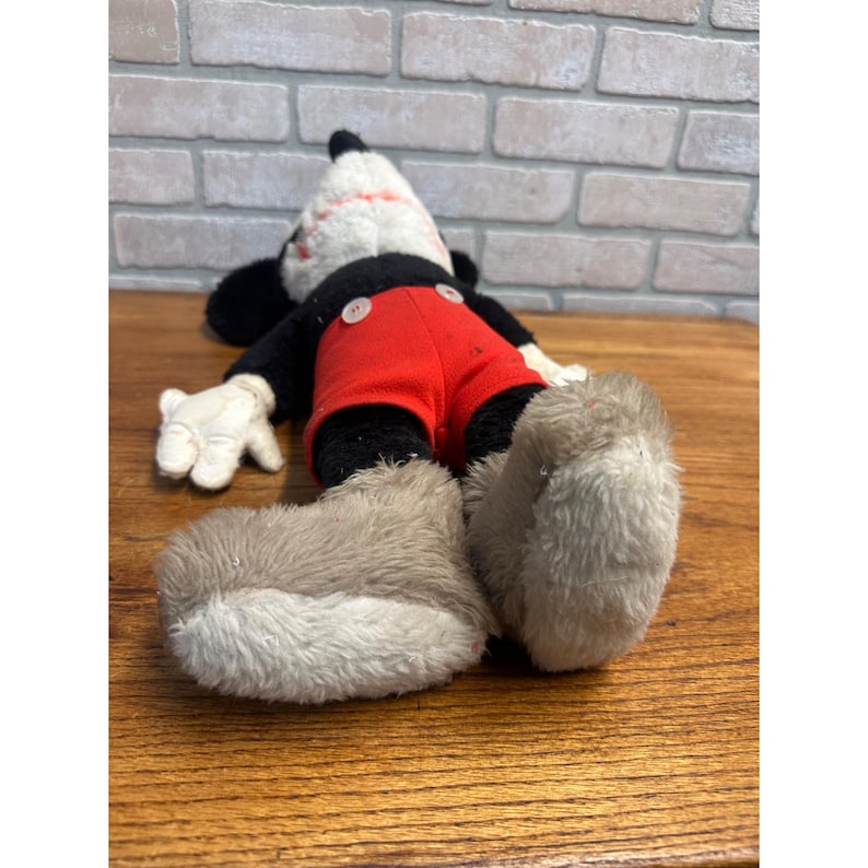 May include: Vintage Mickey Mouse plush toy. The plush has a black body, red shorts, white gloves, and large, fluffy grey shoes. The head is white with black ears and a red and black design. The toy is lying on a wooden surface.
