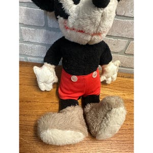 May include: A vintage Mickey Mouse soft toy. The plush has a black torso, red shorts with white buttons, white gloves, and large grey shoes. The face is white with black ears and a red smile. The toy is on a wooden surface.