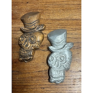 May include: Two skull-shaped ornaments, one finished in a natural wood tone and the other in a metallic silver color. Each skull is depicted wearing a top hat with a curved brim, featuring detailed carved facial features including eye sockets and teeth.
