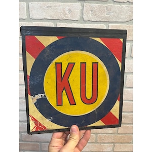 May include: A square metal sign featuring a central yellow circle with the red letters KU inside, surrounded by a dark blue ring and diagonal red and cream stripes. The sign has a black border and shows signs of wear and paint chipping.