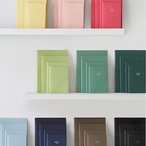 May include: A collection of notebooks arranged in sets of three on white shelves. Each set features a monochromatic color scheme, including yellow, pink, red, light green, sage, dark green, light blue, navy, brown, and black. Each notebook has text.