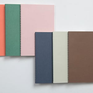 May include: A collection of colorful notebooks in shades of coral, green, pink, navy, cream, and brown. The notebooks are arranged in two stacks, with the pages visible and the binding on the left side.