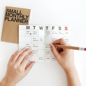 May include: A small monthly planner with a brown cover and a white calendar page. The calendar is open to a week, with a hand holding a pencil writing on it. The text "SMALL MONTHLY PLANNER" is printed on the cover.