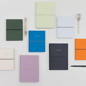 May include: A collection of notebooks in various colours, each with the word "AGENDA" printed on the cover. The notebooks are in shades of green, blue, orange, and purple. A pen and tape are also included.