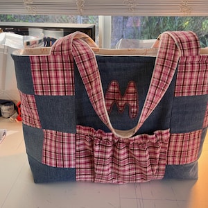 Op de afbeelding: Een patchwork draagtas gemaakt van denim en geruit stof. De tas heeft twee grote zakken aan de voorkant en één zak aan de achterkant. De tas is gevoerd met witte stof en heeft een grote "M" geborduurd op de voorkant.