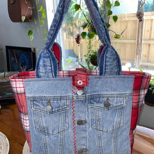 May include: A denim tote bag with a red plaid lining and handles. The front of the bag features a repurposed denim jacket design with pockets and button closures. Red stitching accents the front. The bag is held up by a hand.