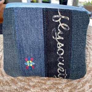 May include: A small denim pouch with a light blue zipper and a colorful embroidered design. The pouch features a vertical black stripe with white cursive text. The denim is a patchwork of different shades of blue.