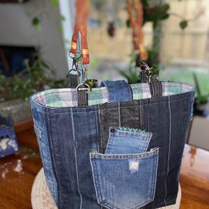 May include: A denim tote bag with a patchwork design. The bag features two front pockets made from repurposed jeans, a plaid interior lining, and sturdy handles with metal clasps. The bag is sitting on a woven placemat.
