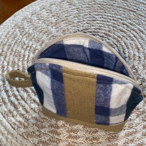 May include: A small, open coin purse with a plaid interior and a tan exterior. The purse has a zipper closure and a small loop handle. The plaid pattern features blue and white squares. The purse is sitting on a woven surface.