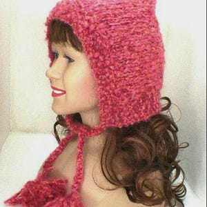 May include: A handmade, textured, pink knit hat with a pointed top and ear flaps. The hat features long ties with matching pink pom-poms. The hat is displayed on a mannequin head.