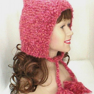 May include: A pink knit hat with a pointed top and ear flaps. The hat has long ties with pom-poms at the ends. The hat is made of a textured, chunky yarn.