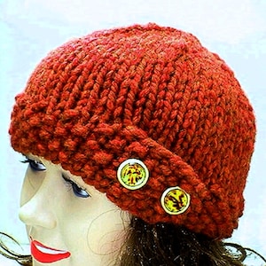May include: A knitted, rust-colored beanie hat with a folded brim. The hat is adorned with two decorative, round buttons with a yellow and green design. The hat is made of a thick, textured yarn.