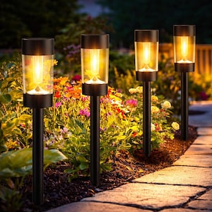May include: Four black solar-powered garden lights with a warm, yellow glow. Each light has a cylindrical glass enclosure and sits atop a black stake. The lights are placed in a garden bed with colorful flowers and a stone pathway.