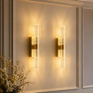 May include: Two wall sconces with a gold finish and clear, textured glass tubes. The sconces are mounted on a light-colored wall, and the design includes gold bands. The sconces are illuminated, casting a warm glow.