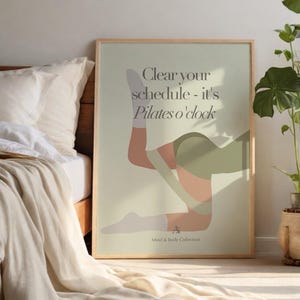 May include: A framed poster with a light green background features a Pilates pose illustration in shades of peach, green, and white. The text "Clear your schedule - it's Pilates o'clock" is displayed at the top. The poster is part of the Mind & Body Collection.