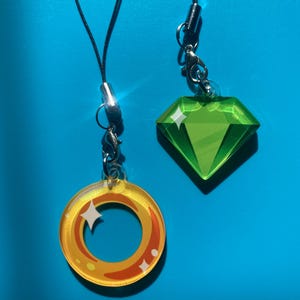 May include: Two phone charms, one is a green diamond shape and the other is a gold ring with a white star inside.