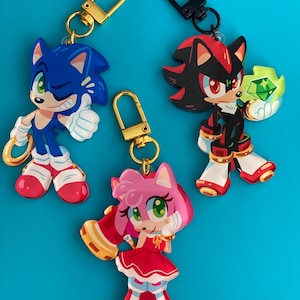 May include: Three character keychains featuring Sonic the Hedgehog, Shadow the Hedgehog, and Amy Rose. The keychains have gold and black hardware. Sonic is blue, Shadow is black and red, and Amy is pink. The background is a solid blue.