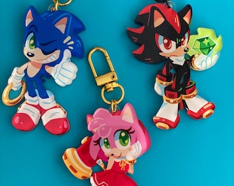 May include: Three character keychains featuring Sonic the Hedgehog, Shadow the Hedgehog, and Amy Rose. The keychains have gold and black hardware. Sonic is blue, Shadow is black and red, and Amy is pink. The background is a solid blue.