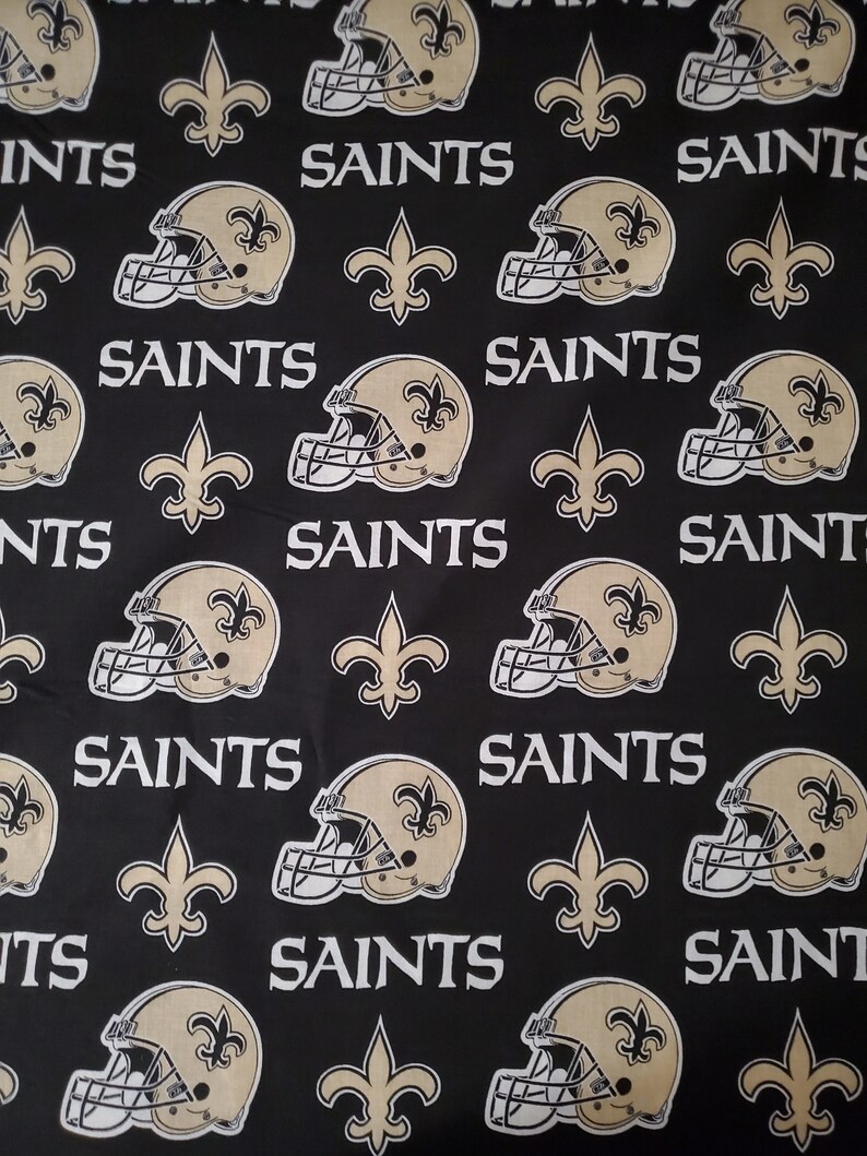 May include: Black fabric with a repeating pattern of gold football helmets, fleur-de-lis, and the word "Saints".
