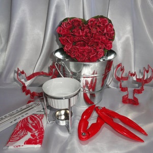 May include: A red heart-shaped arrangement of artificial roses in a silver metal bucket, a white ceramic fondue pot with a metal stand, a red lobster cracker, and a red lobster cookie cutter on a white surface.