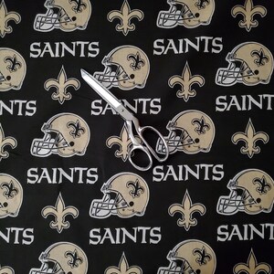May include: Black fabric with a repeating pattern of gold fleur-de-lis, football helmets, and the word "Saints".