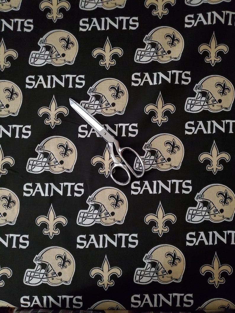 May include: Black fabric with a repeating pattern of gold fleur-de-lis, football helmets, and the word "Saints".