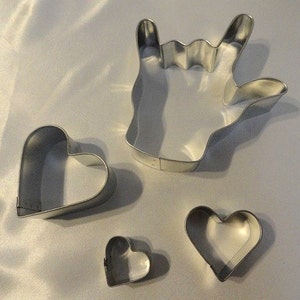 May include: Four metal cookie cutters: a large hand making the sign for 'I love you', a medium-sized heart, and two smaller hearts.