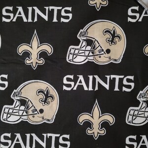 May include: Black fabric with a repeating pattern of gold football helmets, fleur-de-lis, and the word "Saints".