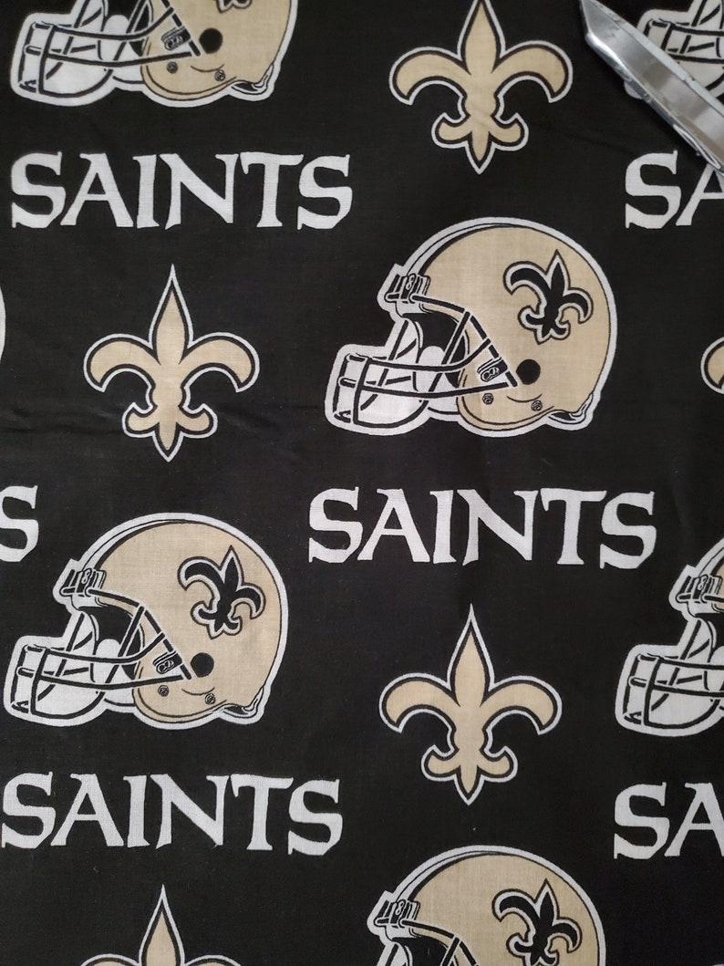 May include: Black fabric with a repeating pattern of gold football helmets, fleur-de-lis, and the word "Saints".