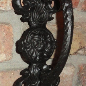 May include: Black cast iron door knocker with a decorative design featuring a floral motif and a stylized face. The knocker has a loop handle for knocking.