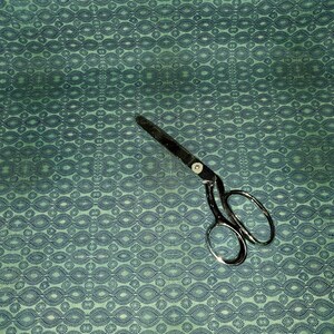 May include: A teal fabric with a repeating pattern of dark blue circles and ovals. A pair of silver scissors is lying on the fabric.