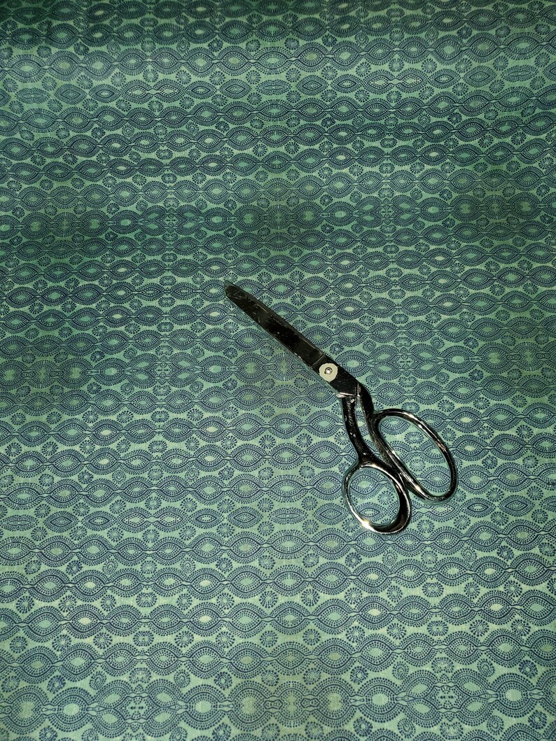 May include: A teal fabric with a repeating pattern of dark blue circles and ovals. A pair of silver scissors is lying on the fabric.
