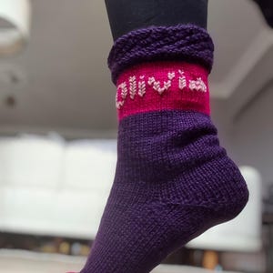 May include: Purple and pink knitted socks with a pink band and the word "Olivia" embroidered in white on the band.