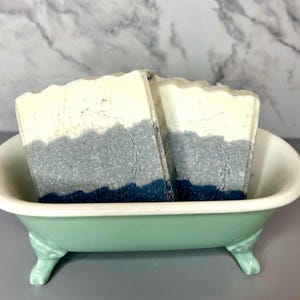 Puede incluir: Dos jabones artesanales con un diseño en capas en un pequeño jabonero en forma de bañera de color verde menta. Los jabones tienen una capa superior blanca, una capa media gris y una capa inferior azul oscuro. El jabonero tiene patas decorativas.