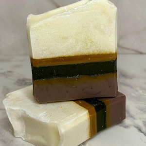 May include: Two handmade soap bars. The top bar is stacked on the bottom bar. The soaps have a layered design with a creamy white top, followed by a brown, green, and dark brown layer. The bottom bar has a creamy white top and a layered design.