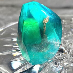 May include: A faceted, gemstone-shaped object with a translucent teal and green color scheme. Gold-colored inclusions are visible within the gem. The object is displayed on a clear, patterned surface.