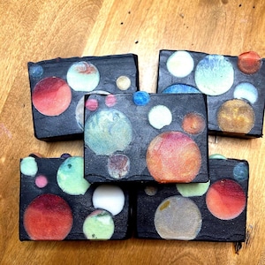 Puede incluir: Cinco pastillas de jabón rectangulares con un fondo negro y diseños de planetas coloridos. Los planetas son de varios tamaños y colores, incluyendo rojo, naranja, azul y blanco. Las pastillas de jabón están dispuestas sobre una superficie de madera.