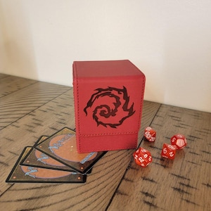 May include: A red card storage box with a black graphic design, next to several red dice and a few trading cards. The box is square and appears to be made of textured material. The dice are white with red numbers.