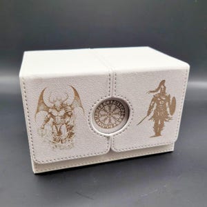 May include: White leather card box with a decorative design featuring a mythical creature, a warrior, and a circular symbol. The box is closed.