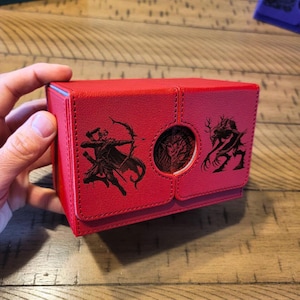 May include: Red leather deck box with a black and white illustration of a person with a bow and arrow and a creature with antlers on the front.