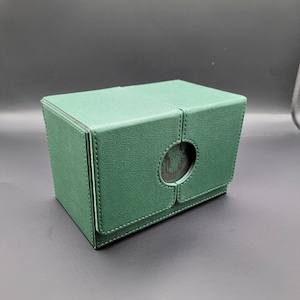 May include: A green leather deck box with a circular window on the front. The box is closed and has a stitched seam down the middle.