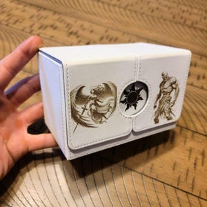 May include: White card deck box with a gold design featuring a winged figure, a sun, and a warrior. The box has a hinged lid and a small window.