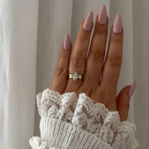 Peut inclure: Une bague en or avec une pierre centrale en diamant taille marquise. La bague présente un design de bande détaillé. La main montre des ongles longs en forme d'amande peints en rose clair. Une manche de pull blanc avec une garniture en dentelle est visible.