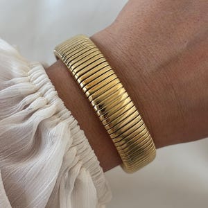 Peut inclure: Un bracelet en métal doré composé d'une série de maillons rectangulaires lisses et connectés formant une bande flexible et continue. La surface du métal présente une finition polie et réfléchissante, créant un aspect uniforme sur la pièce.