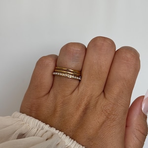 Könnte beinhalten: Ein Set aus drei gestapelten Ringen an einem Finger. Der oberste Ring ist ein schlichter Goldring, der mittlere Ring hat ein strukturiertes, gedrehtes Golddesign und der untere Ring ist ein dünner Goldring mit einer Reihe kleiner, weißer Perlen.