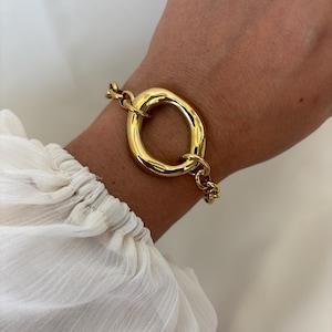 Peut inclure: Un bracelet doré composé d'un grand maillon central ovale et lisse relié à une chaîne classique. La surface métallique polie reflète la lumière, créant un aspect brillant et métallique qui entoure la zone du poignet.