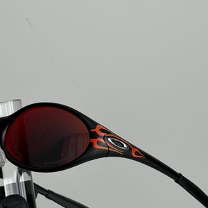 May include: Black sunglasses with red lenses and a flame design in orange and yellow on the temples. The sunglasses have a sleek, modern design.