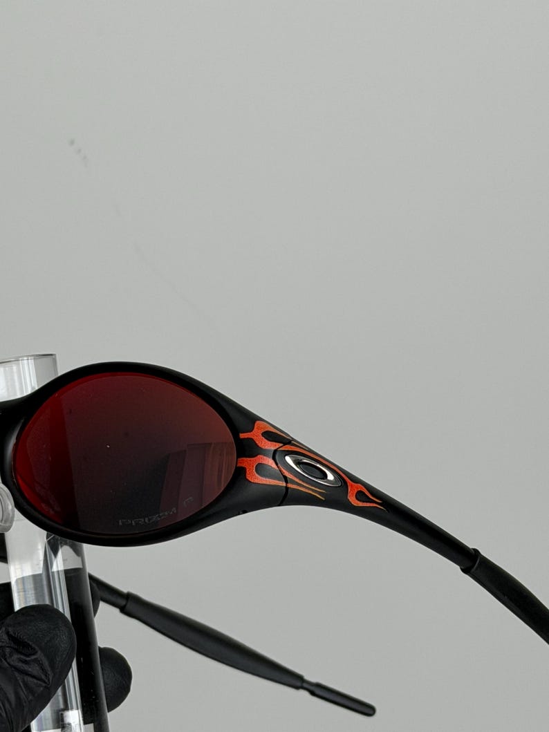 May include: Black sunglasses with red lenses and a flame design in orange and yellow on the temples. The sunglasses have a sleek, modern design.